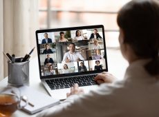 View over businesslady shoulder seated at workplace desk look at computer screen where collage of many diverse people involved at video conference negotiations activity, modern app tech usage concept